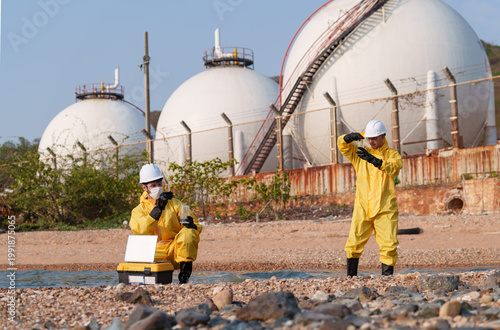 Workers collect water samples near industrial storage tanks in a coastal area during daylight hours