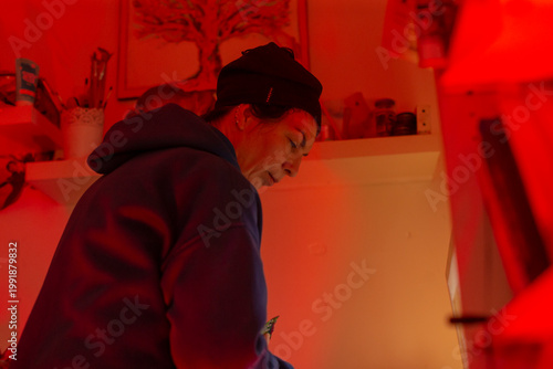 woman in red lit studio editing images on phone, focused expression, shelves with frames, desk lamp casting warm shadow, intimate closeup, hoodie and beanie, creative concentration