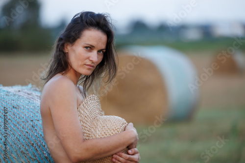 Young woman in countryside holding straw hat, natural summer portrait with hay bales
