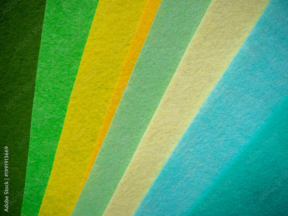Fototapeta premium Multiple colorful felt fabric sheets fanned out diagonally displaying a nature-inspired color spectrum — dark forest green, bright green, yellow-green, golden amber, pale mint, cream/beige, light blue