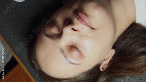 Woman lying down on a treatment bed, receiving a cosmetic eyebrow procedure, with a beautician's hands gently cleaning and shaping her brows using cotton during a facial care session