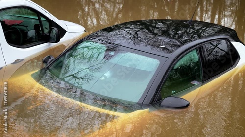 Disaster flooding submerging passenger cars in deep muddy water. Storm runoff rising and trapping vehicles during severe weather event. Climate emergency escalating and causing widespread transport