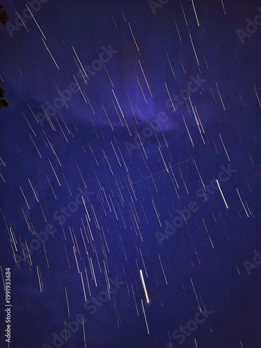 Quadro su tela A long-exposure photo captures streaks of stars moving across the night sky, with a striking red nebula glowing vividly in the center, creating a cosmic and mesmerizing scene