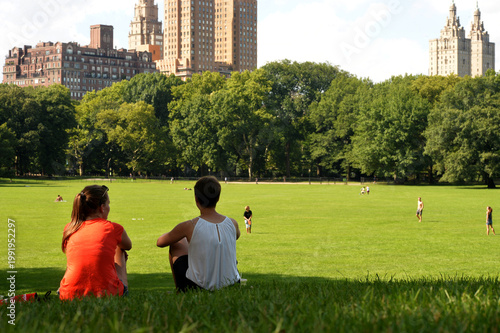 Two Companions Resting On Grass Observing Cityscape And Jogging Passersby While Conversing new york