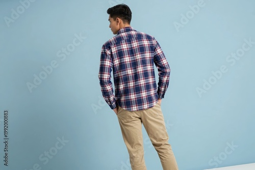 Man stands facing away in a checkered shirt and khaki pants against a light blue wall
