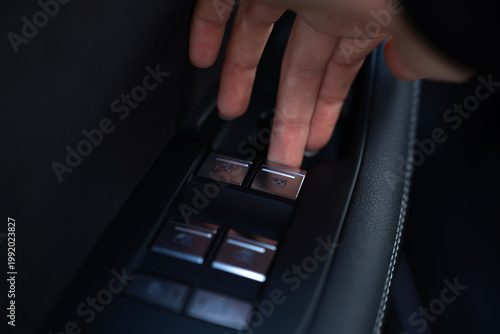 Foto Close-up of fingers pressing power window switch on driver door panel inside modern car, with other window controls and mirror knob nearby
