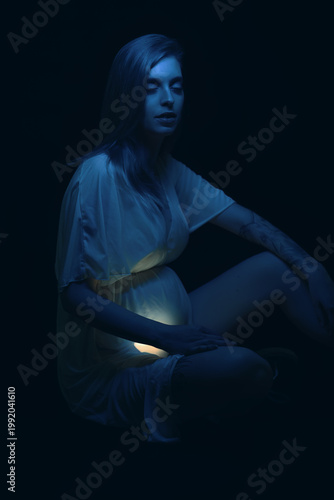 Low key photo of sad young pregnant woman illuminated by blue fluorescent light is sitting and holding her belly. Vertical. 