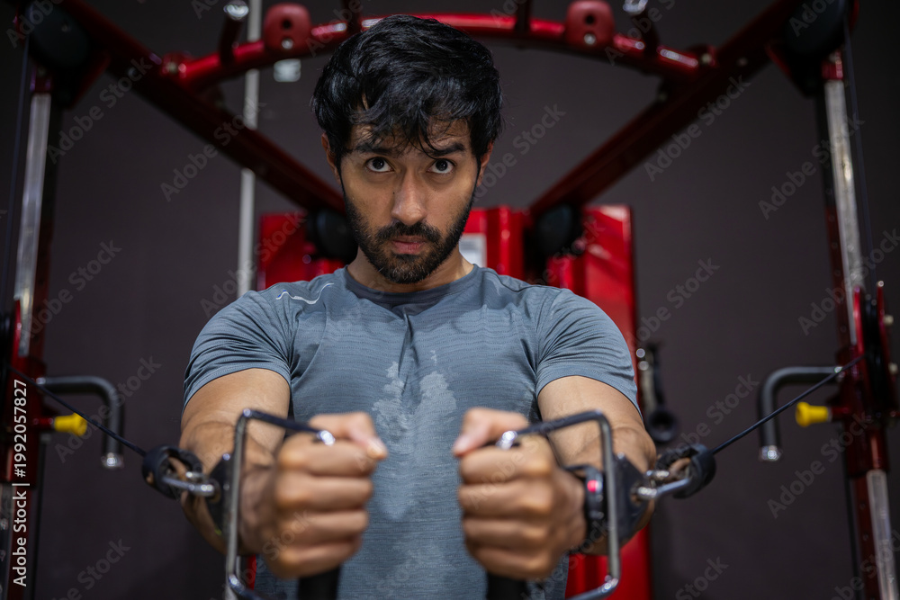 Fototapeta premium Young adult man doing intense workout at gym using cable machine displaying deep determination and focused emotional expression while building physical strength during fitness exercise