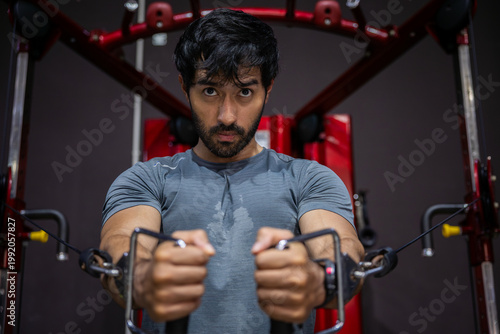Young adult man doing intense workout at gym using cable machine displaying deep determination and focused emotional expression while building physical strength during fitness exercise