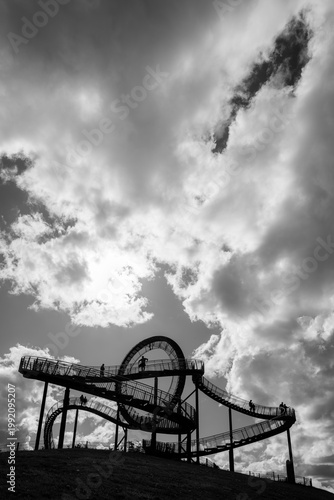 A silhouette of a popular observation deck in Duisburg on the Rhine (Germany), featuring a few people backlit against impressive fair-weather clouds. The steel structure is located on a slag heap. 