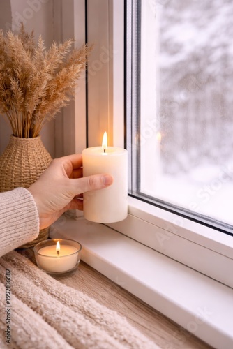 Hand holds a lit candle beside a snowy window, illustrating indoor warmth, winter comfort, and possible draft inspection.