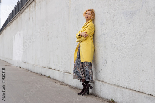 Thoughtful woman in yellow coat leaning against white concrete wall outdoors