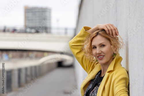 Smiling mature woman touching curly hair while posing outdoors in yellow coat