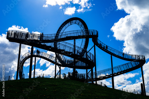 A silhouette of a popular observation deck in Duisburg on the Rhine (Germany), featuring a few people backlit against impressive fair-weather clouds and clear blue sky. 