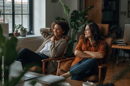 Office colleagues sharing quiet break conversation in sunlit workspace with plants and warmth