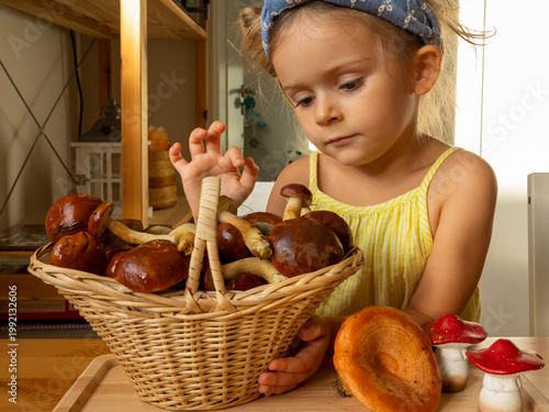 A curious cute toddler girl sits next to a wicker basket filled with Bay bolete wild mushrooms