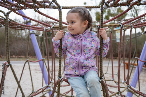 Young smiling girl in purple fleece jacket holds the thick ropes of a climbing net at outdoor playground