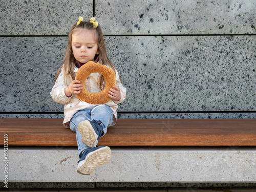 Cute toddler girl with long hair and yellow bows sits on a wooden bench holding a large sesame bagel
