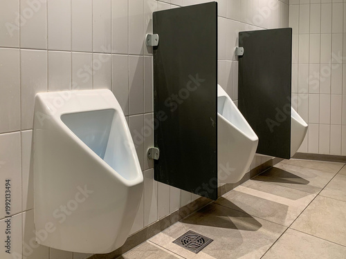 White ceramic urinals with black privacy dividers lines a clean tiled public restroom WC wall