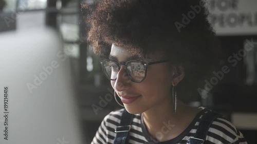 Close-up portrait of a focused young woman with afro hairstyle and glasses looking at computer screen in a modern interior. Stylish casual outfit, soft natural light and calm concentrated expression