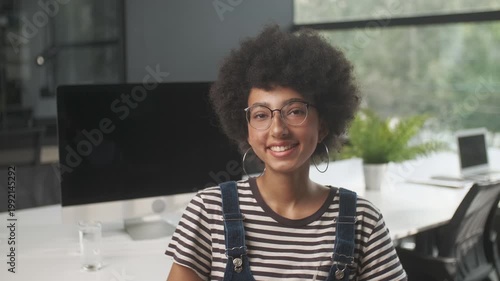 Portrait of a smiling young woman with afro hairstyle and eyeglasses looking at camera in a modern office interior. Friendly female professional in striped shirt and denim overalls posing at workplace