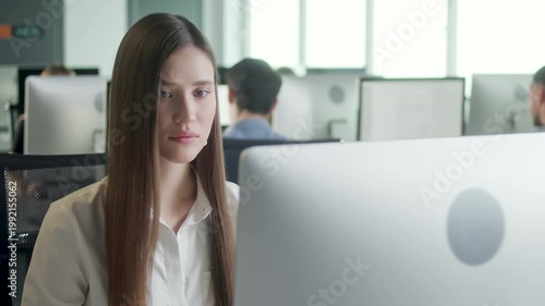 Young businesswoman working at desktop computer in a modern open plan office. Concentrated female office employee looks at monitor while sitting at her workplace, with coworkers and office desks
