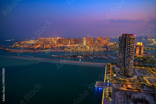 Aerial night view of Doha, Qatar, featuring illuminated skyscrapers, marina, and waterfront developments under a twilight sky along the Arabian Gulf