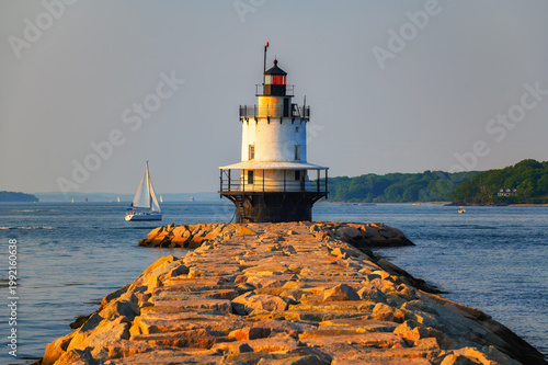 Spring Point Ledge Lighthouse at the end of a granite breakwater with sailboat on Casco Bay in warm golden hour light in South Portland, Maine, USA