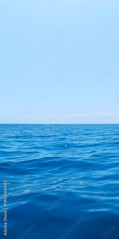 Fototapeta premium Vast and serene expanse of deep blue ocean water stretches calmly towards the horizon under a clear, pale blue sky on a bright sunny day.