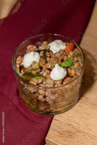 Close-up of a gourmet farro salad with vegetables and creamy cheese in a glass, served on a rustic wooden table with warm, elegant presentation.