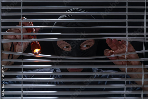 A burglar checks if there are people inside a house by peering through the slats of a Venetian blind with a flashlight