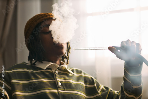 Quadro su tela Black teenager exhaling vapor from hookah hose indoors, illustrating youth smoking habit and nicotine use