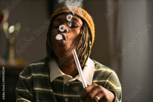 Quadro em tela Young Black man exhaling smoke rings while holding smoking device indoors, illustrating cannabis use and substance habit