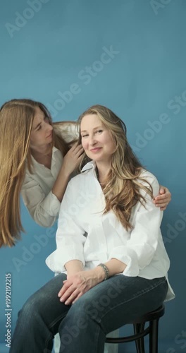 Vertical video of beautiful mature mother and adult daughter in white shirts and jeans in the photostudio on blue paper background. Two female generations. Mother's day concept