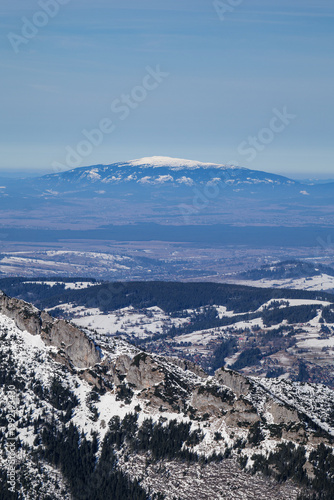 Snow-covered mountain peak stands above valley during winter with clear blue sky overhead.View from Kasprowy Peak in Zakopane. Long focal length. Babia Góra.