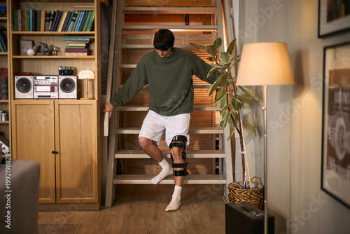 Young adult man with disability descending stairs while wearing knee brace at home, managing mobility recovery. Useful for rehabilitation, accessibility, injury recovery campaigns, wide shot