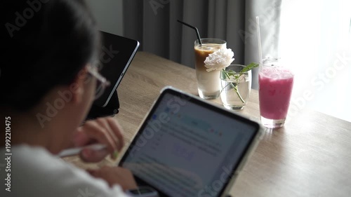 Over the shoulder view of a student girl using a digital tablet for online learning at a desk with iced latte, pink milk, and a flower, home cafe lifestyle concept