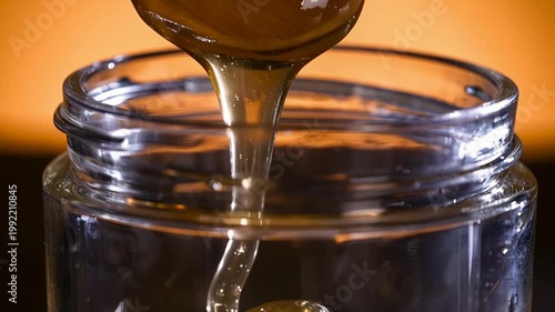 Golden liquid honey pouring from a spoon into a transparent glass jar