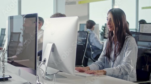 Young businesswoman working at desktop computer in a modern open plan office. Focused female employee sits at her workstation and handles daily tasks while colleagues work at nearby desks in the