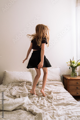 Girl jumping on bed. Joyful child jumping on a bed with messy blankets, hair flying, and sunlight filling the room. A playful, carefree moment of childhood energy and freedom.