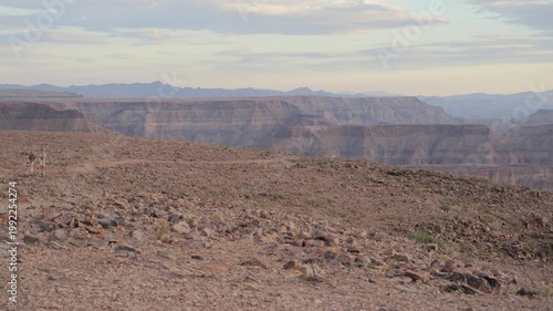 Tourist couple walk through vast desert landscape in Namibia, Fish River Canyon background