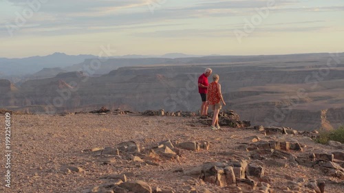 Aged European couple walk to cliff edge, take photographs of Fish River canyon in sunset light