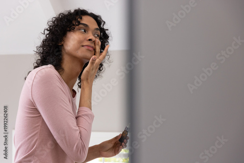 Non-binary adult in pale pink top applying skincare at vanity holding cosmetic bottle near mirror