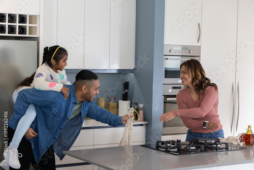 Indian family father carrying child on back passing mesh bag across kitchen island with gas cooktop