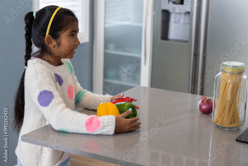 Indian child girl standing at kitchen counter holding peppers, wearing yellow headband, copy space