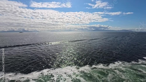 August 13, 2024 - Tracy Arm, USA - View of the ocean with mountains in the distance with a boat creating waves. View of the sky with cloudy conditions.