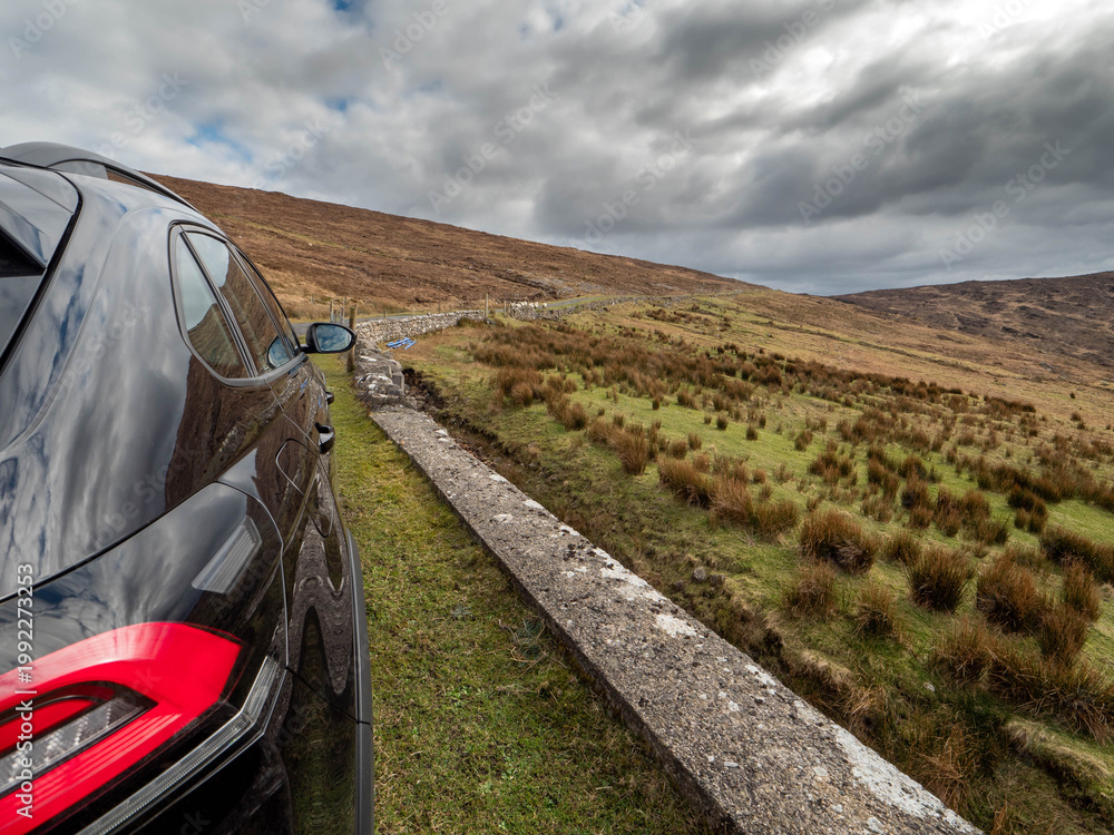 Fototapeta premium Dark SUV parked beside winding rural road. Overcast sky looms above rolling green hills. Vehicle reflects misty, moody atmosphere. Path stretches into distance, inviting exploration Travel concept