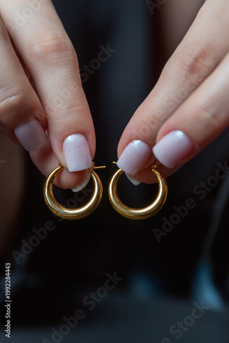Elegant gold hoop earrings held between manicured fingers, soft nail polish, minimalist jewelry shot