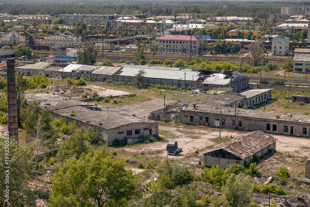 Obraz premium High-angle panoramic view of an industrial zone with dilapidated warehouses, a tall brick chimney, and complex railway tracks leading toward a residential town under a hazy sky.