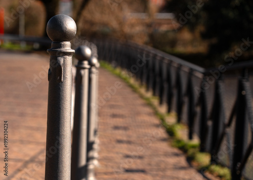 In the foreground, the first in a row of metal posts with spherical tops; in the blurred background, geometric lines created by a railing and its shadow on the paving